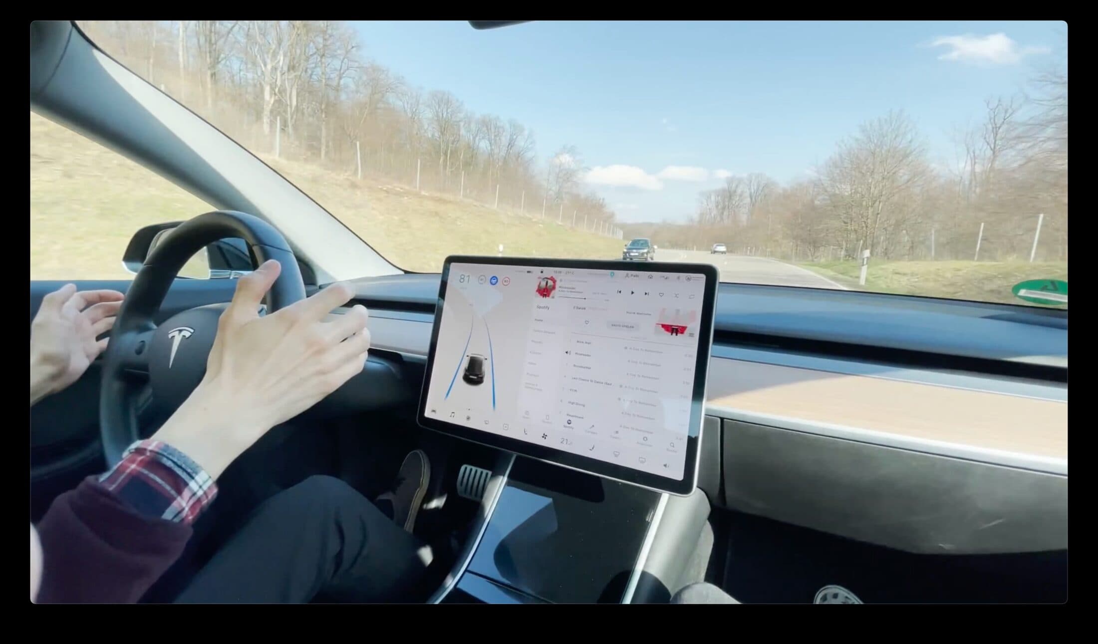 Hands resting on the steering wheel of a Tesla Model 3 on a country road, the centre screen showing the navigation map